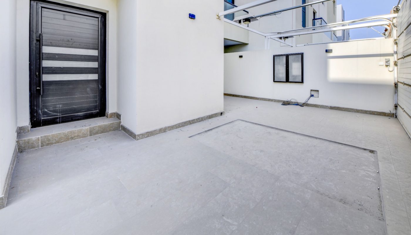 Modern house exterior with a black door, white walls, tiled courtyard, and overhead pipes, taken on a clear day.