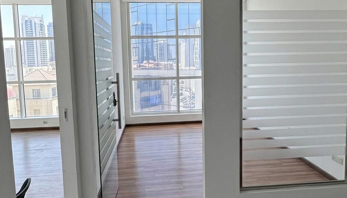 Modern office with glass door and large windows, wood flooring, white walls, and a cityscape visible outside.