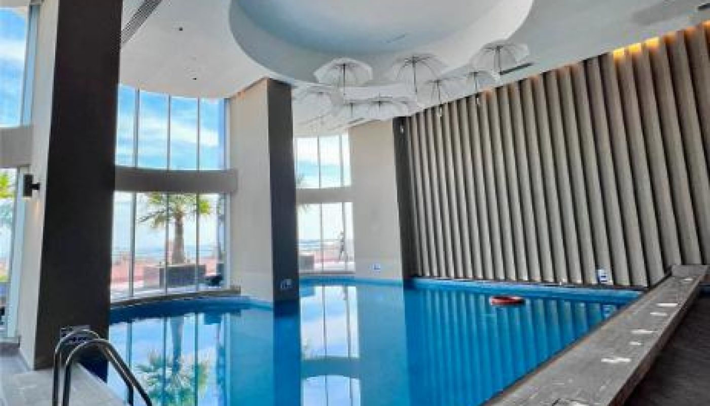 Indoor swimming pool with large windows, high ceilings, and modern decor. Several umbrellas hang upside down from the ceiling as part of the design.