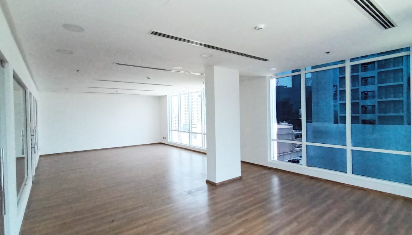 Large empty room with wood flooring, white walls, floor-to-ceiling windows, and ceiling lights, in a modern building with city views visible through the windows.