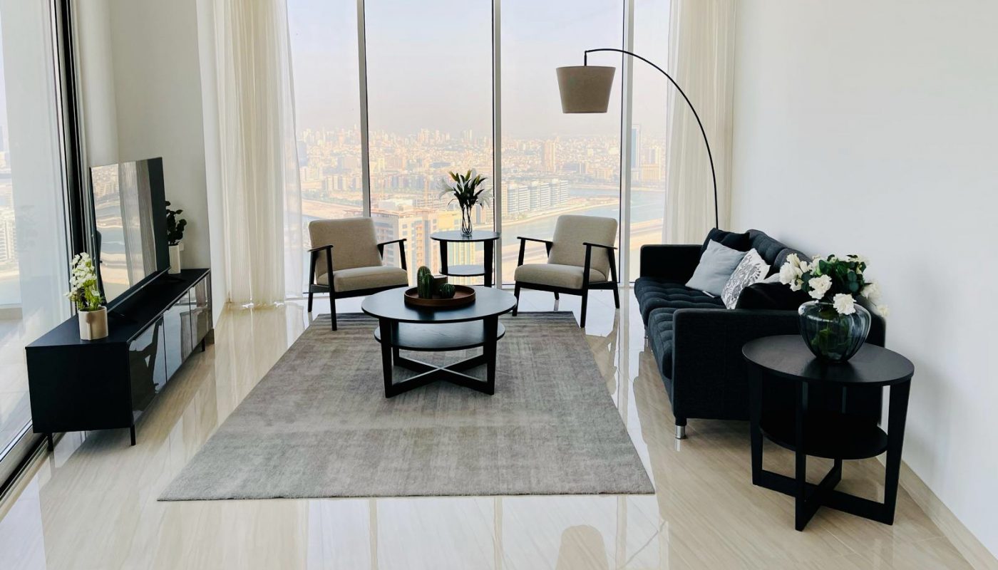 Modern living room with large windows, city view, gray sofa, coffee table, floor lamp, TV stand, and minimalistic decor on glossy tile floor.