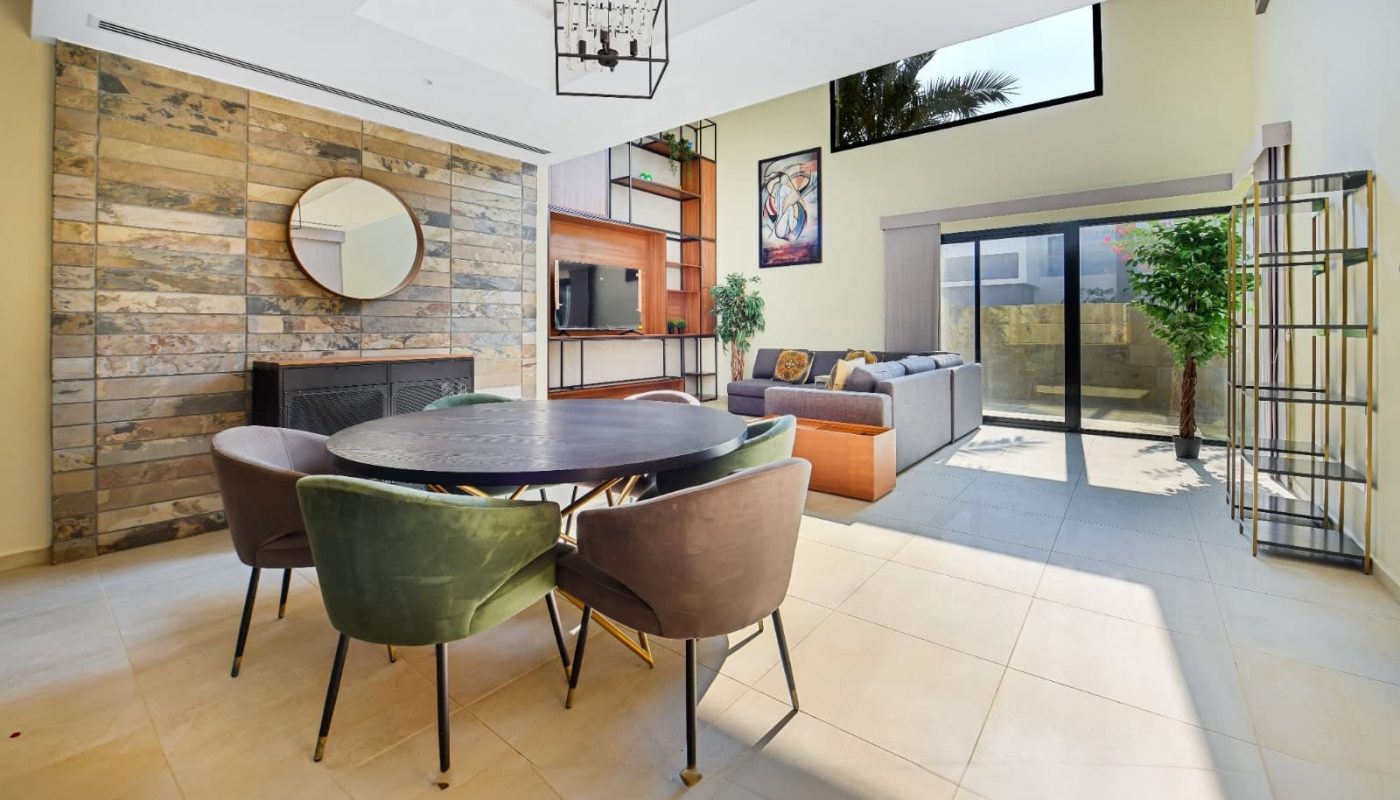 Modern open-plan living area with round dining table, chairs, and a lounge. Stone feature wall, large windows, and bright natural light.