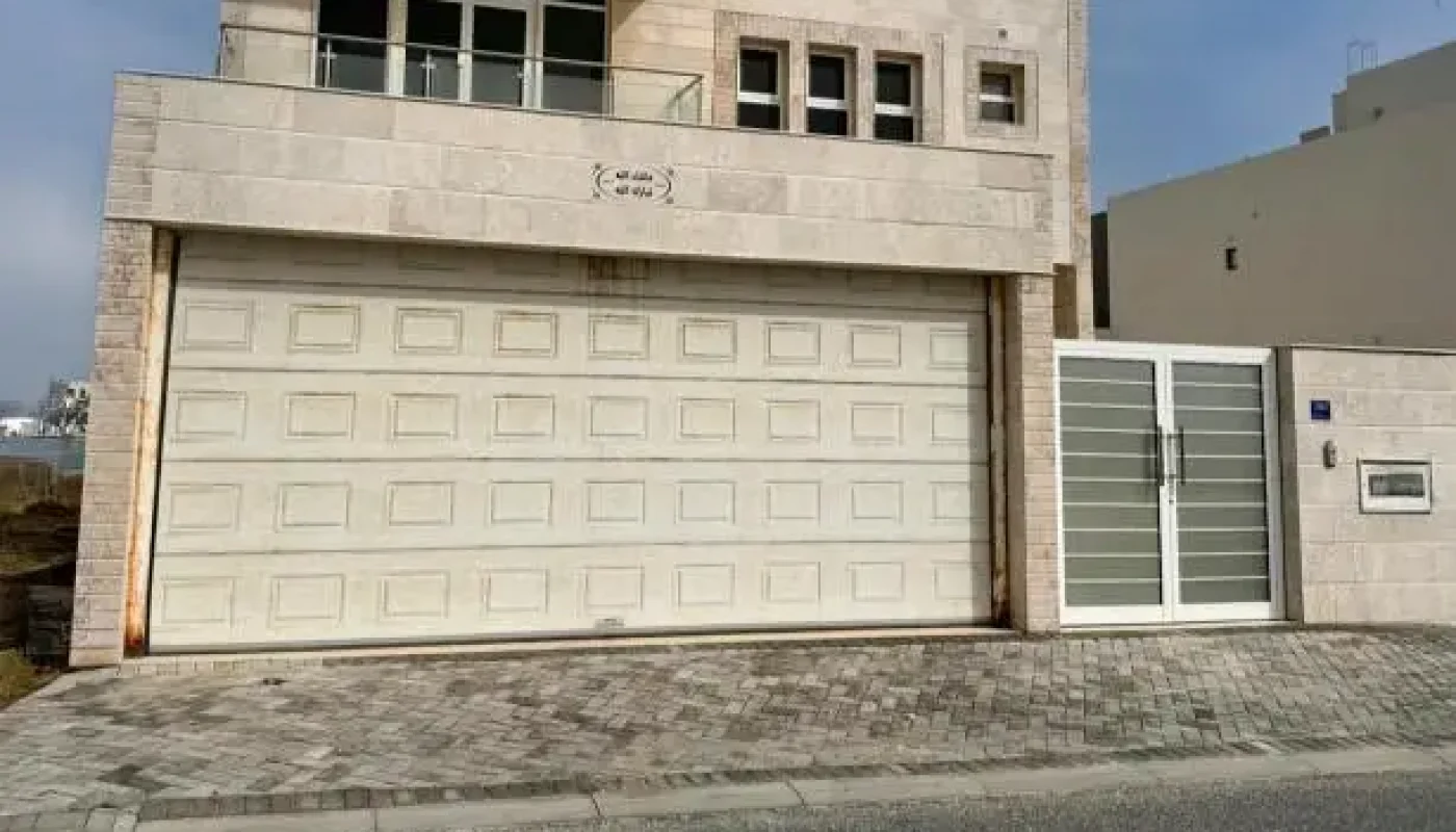 Two-story modern house with light stone exterior, large garage door, metal gate, and small windows, situated along a paved street under a partly cloudy sky.