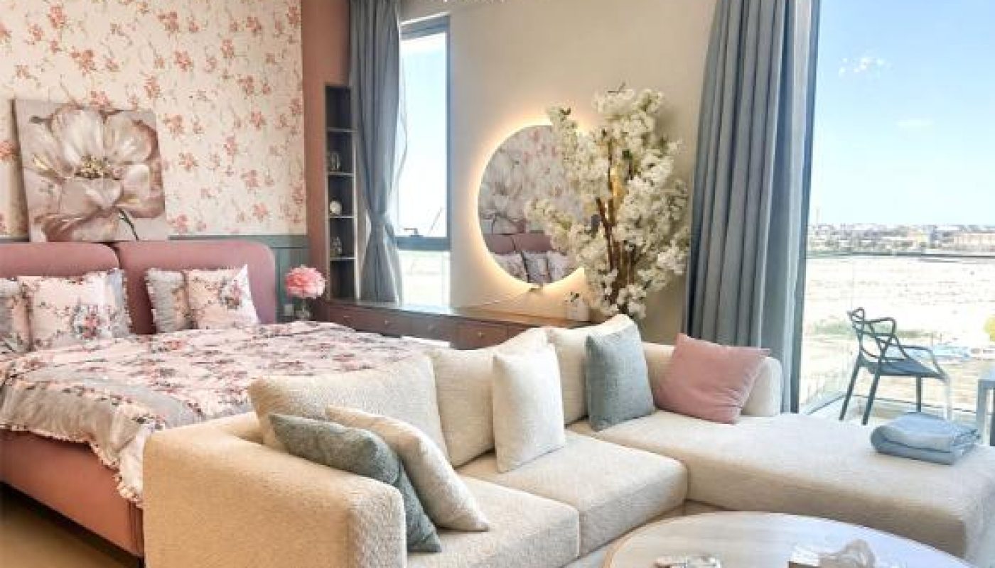A modern bedroom features a floral accent wall, a large bed, a beige sectional sofa, a round wooden table, a chandelier, and floor-to-ceiling windows with city views.