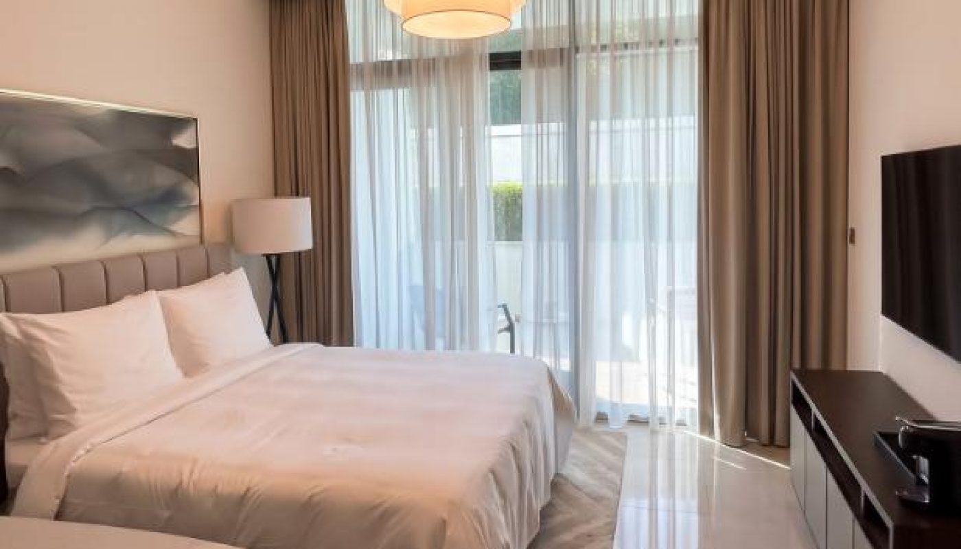 Modern hotel room with a neatly made bed, large window with curtains, a TV on the wall, and a floor lamp beside the bed.