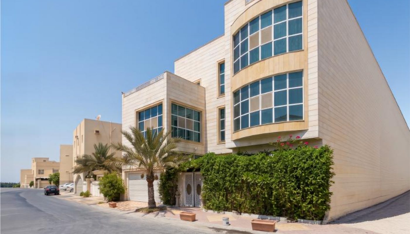 Modern beige building with large windows and green plants in front, located on a quiet, sunny street with a clear blue sky above.