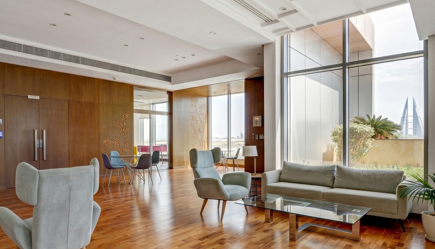 Modern lounge area with wooden flooring, contemporary chairs, a sofa, glass coffee table, and large windows providing natural light and city views.