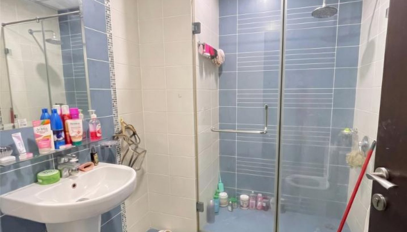 Modern bathroom with blue and white tiles, a wall-mounted sink, mirror with toiletries, toilet, glass shower area, and cleaning supplies visible.