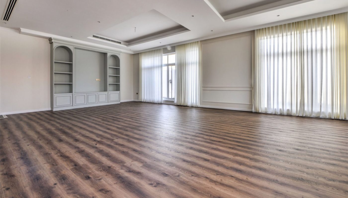 Spacious empty room with wood flooring, large windows covered by sheer curtains, built-in shelves, and recessed ceiling lighting.