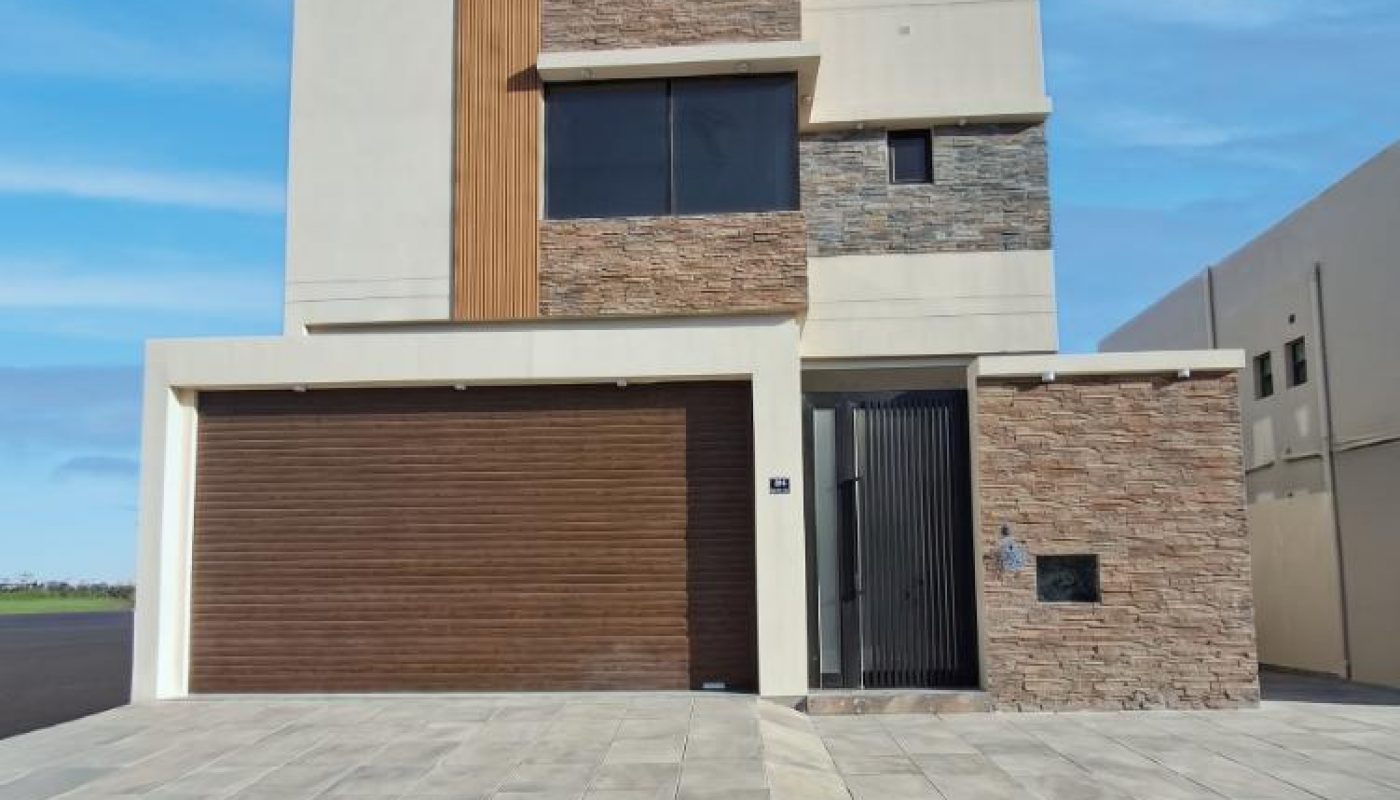 Modern two-story house with a flat roof, brown garage door, stone and wood accents, large paved driveway, and minimalistic design.