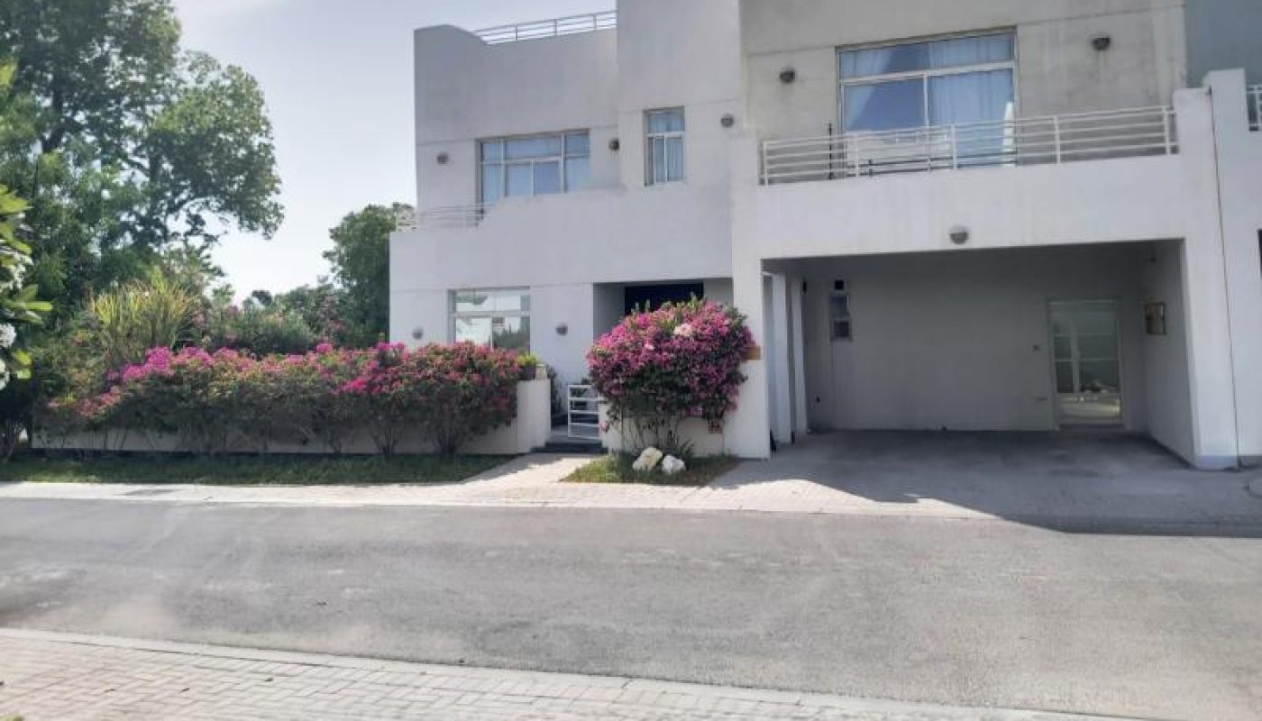 A modern two-story white building with large windows, a balcony, and pink flowering bushes in front, situated on a paved street under a partly cloudy sky.
