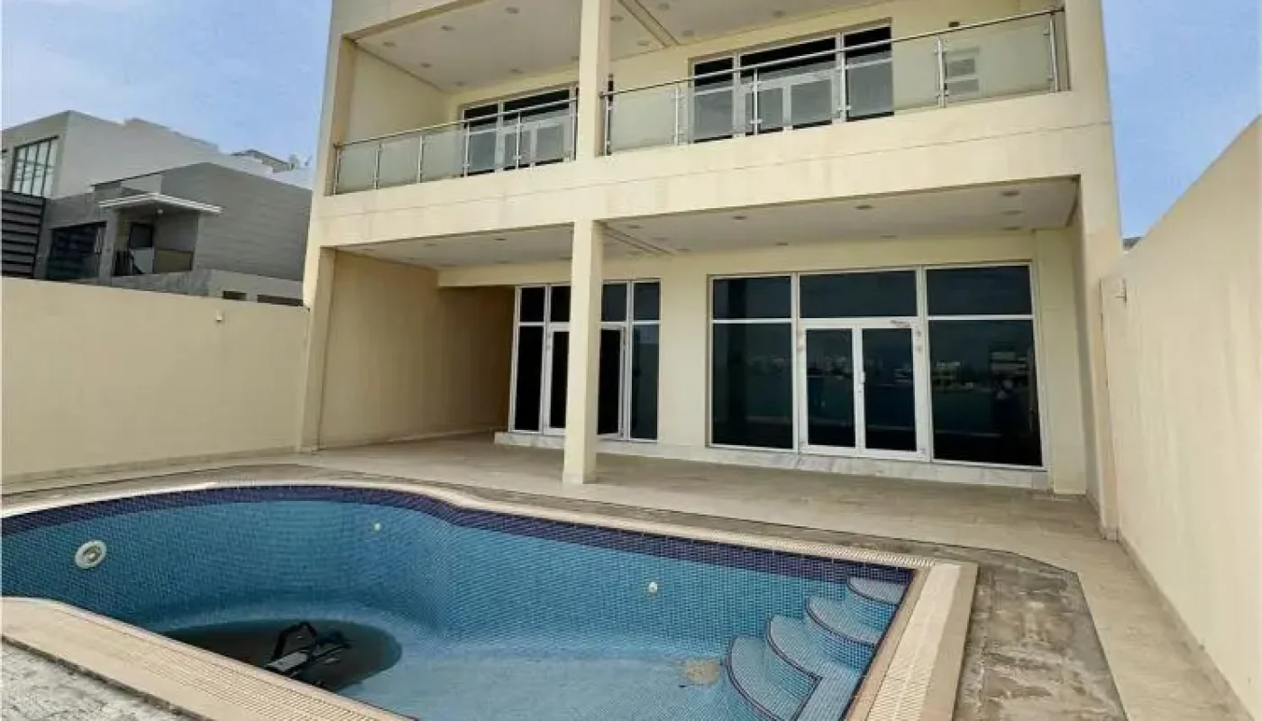 A three-story modern house with large windows and balconies, featuring an empty, unfinished swimming pool in the backyard under a partly cloudy sky.