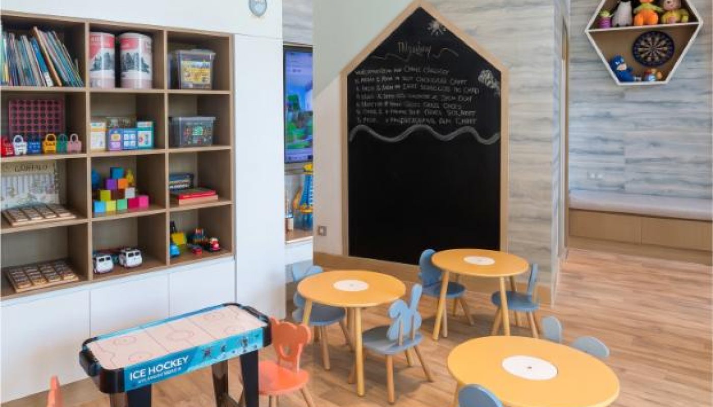 Children’s playroom with round tables, toy storage shelves, a foosball table, ice hockey game, wall chalkboard, books, and hexagonal wall shelves holding toys and a dartboard.