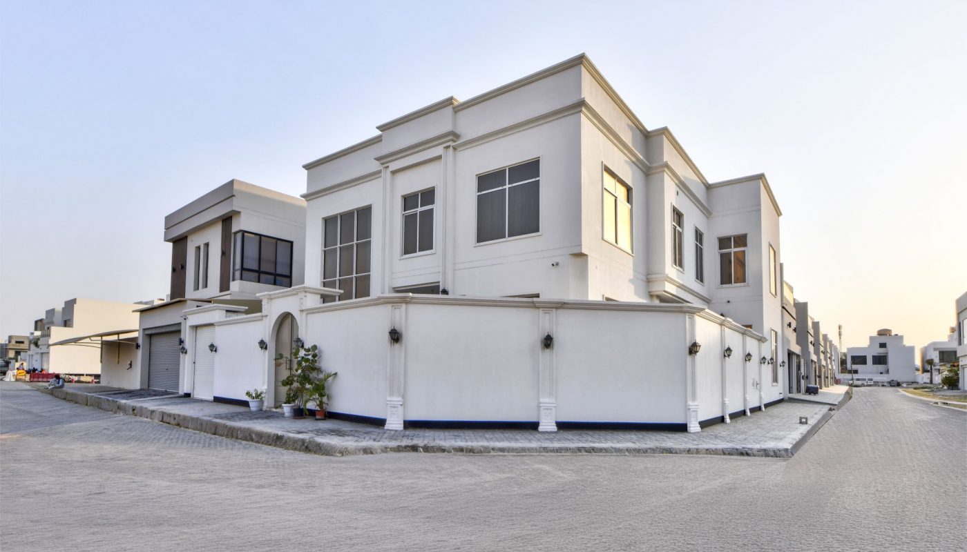 A modern two-story white house with large windows sits on a corner lot in a residential neighborhood under a clear sky.