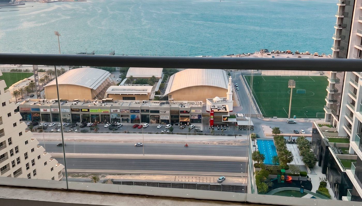 View from a high balcony showing a road, sports fields, buildings, and a large body of water with a distant cityscape under a clear sky.