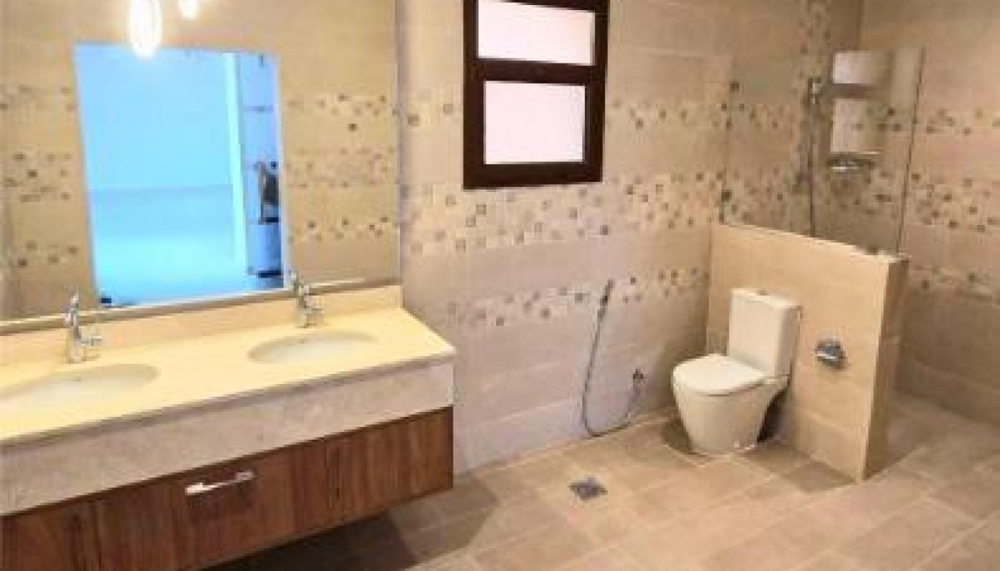 Spacious bathroom with double sink, large mirror, hanging lights, toilet, wall-mounted bidet spray, and beige tile walls and floor.