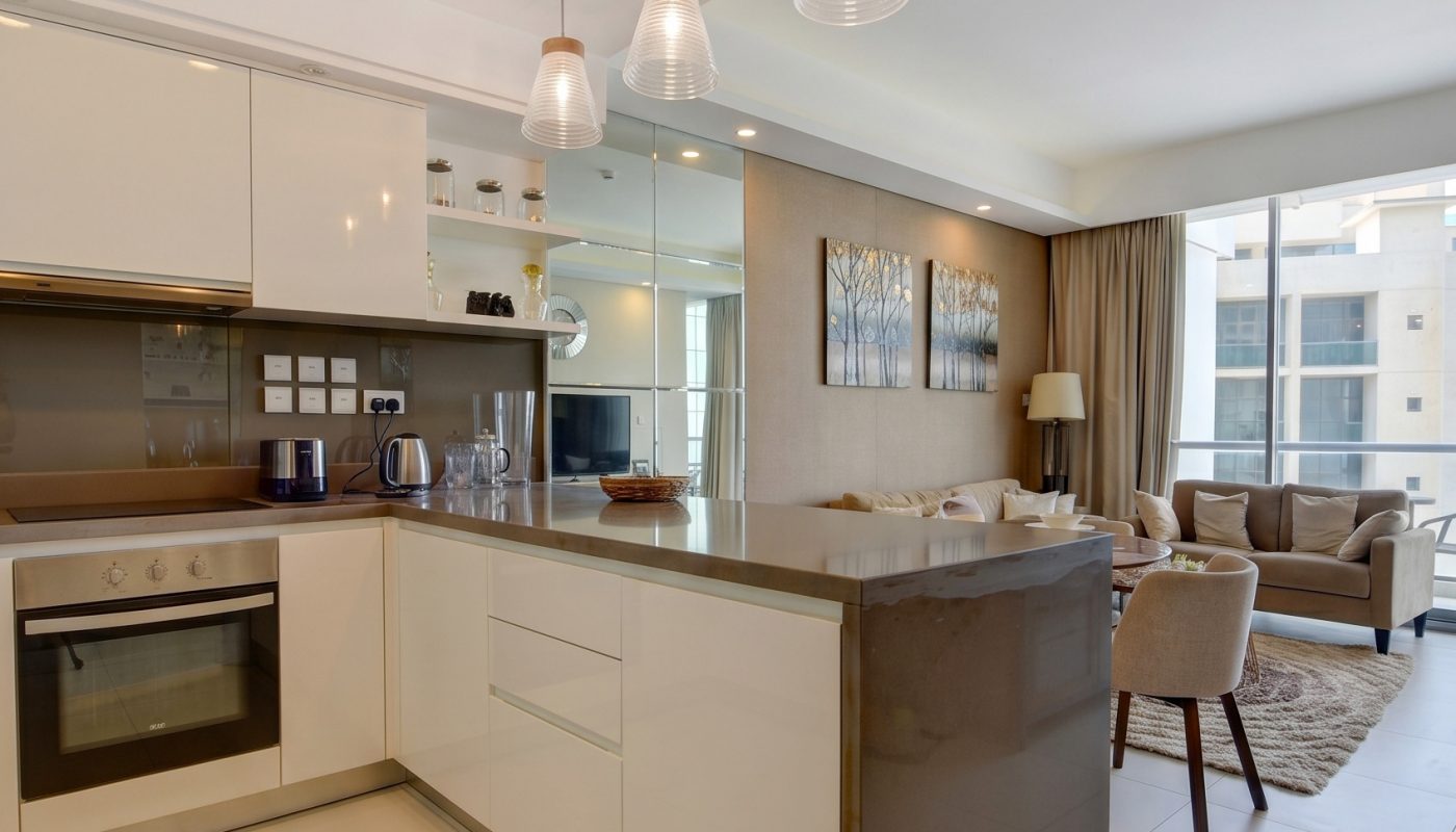 Modern open-plan kitchen and living area with white cabinets, built-in oven, pendant lights, a brown countertop, and a sitting area near large windows.