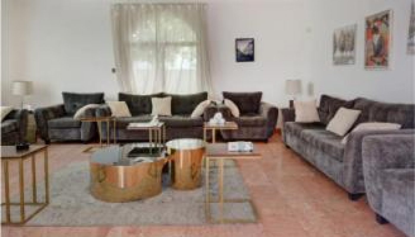 Spacious living room with gray sofas, gold accent tables, lamps, wall art, a chandelier, and large window with sheer curtains. Pink marble flooring is visible in the foreground.