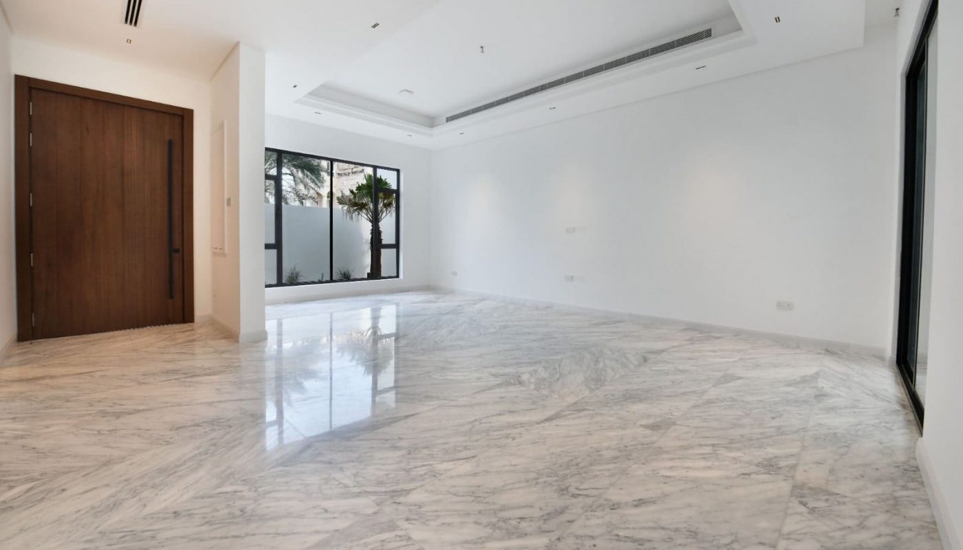 Spacious empty room with marble flooring, white walls, a large wooden door, and windows offering natural light.