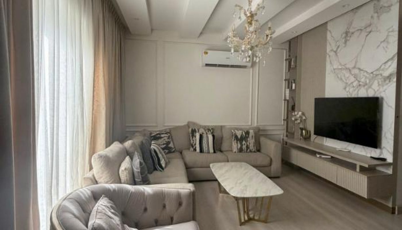 A modern living room with beige sofas, a marble coffee table, a TV on a marble-accented wall, a chandelier, and floor-to-ceiling curtains.