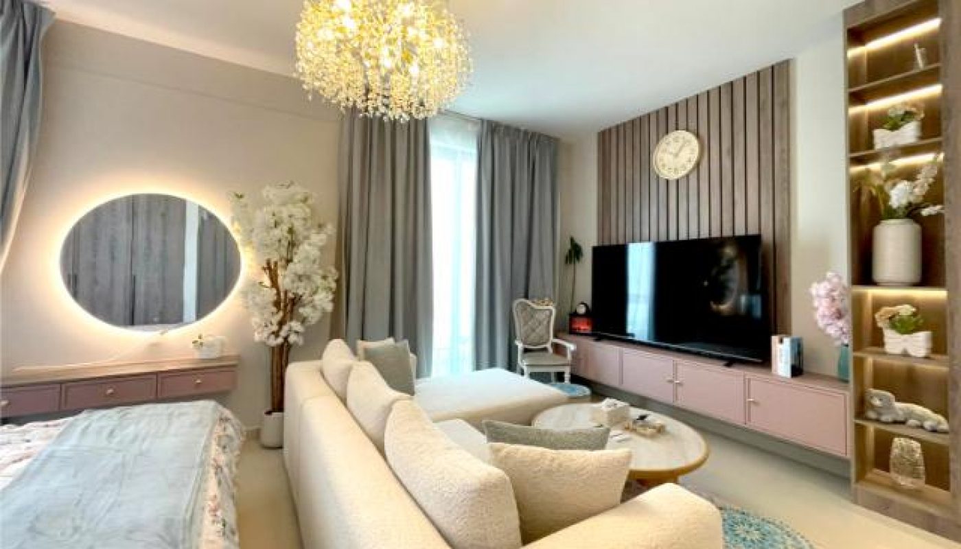Modern living room with neutral decor, a white sofa, large TV, round mirror with backlighting, chandelier, built-in shelves with plants, and floor-to-ceiling curtains.
