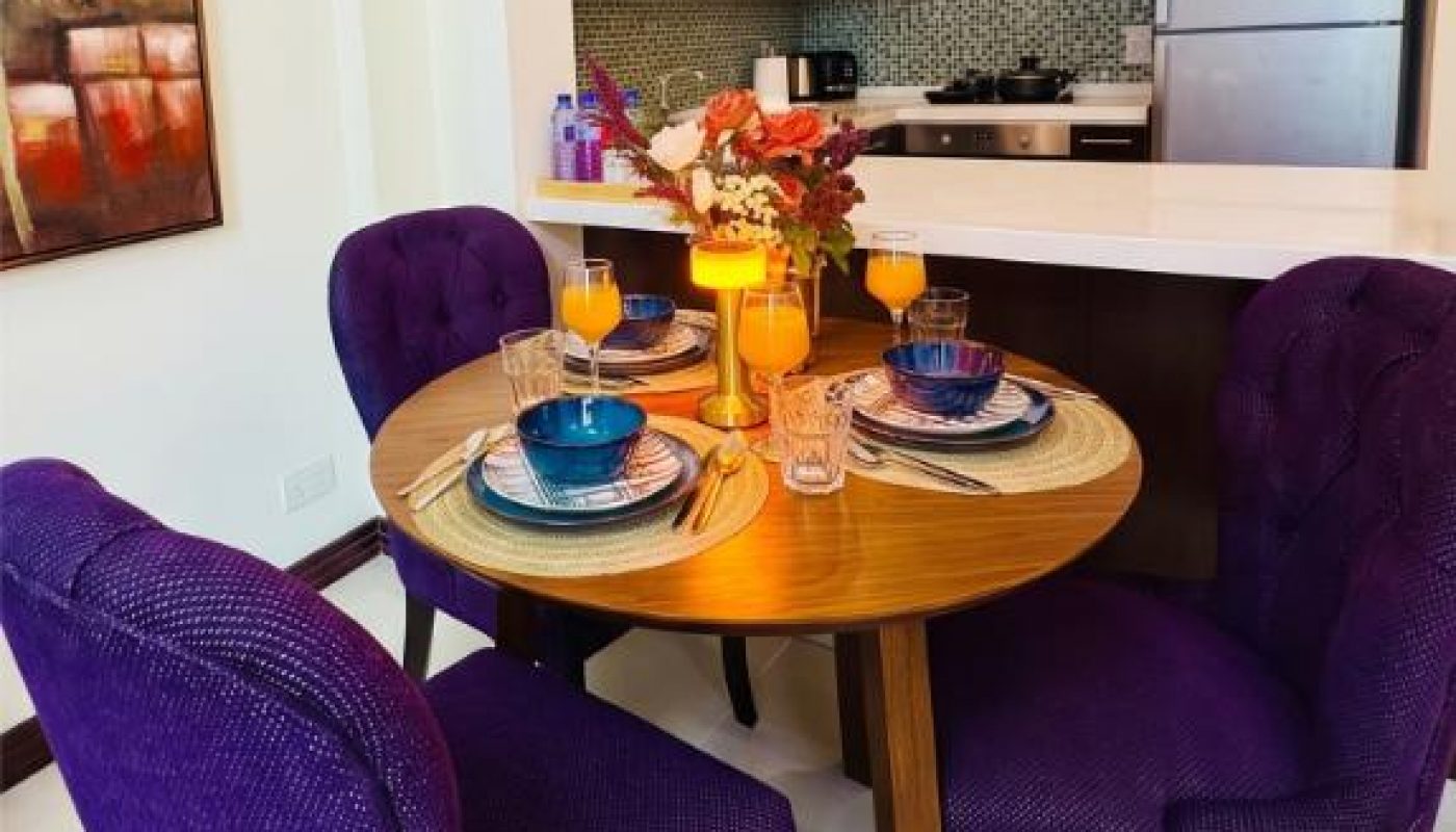 A round wooden dining table set for four with blue and purple chairs, plates, and glasses of orange juice in a modern kitchen-dining area.