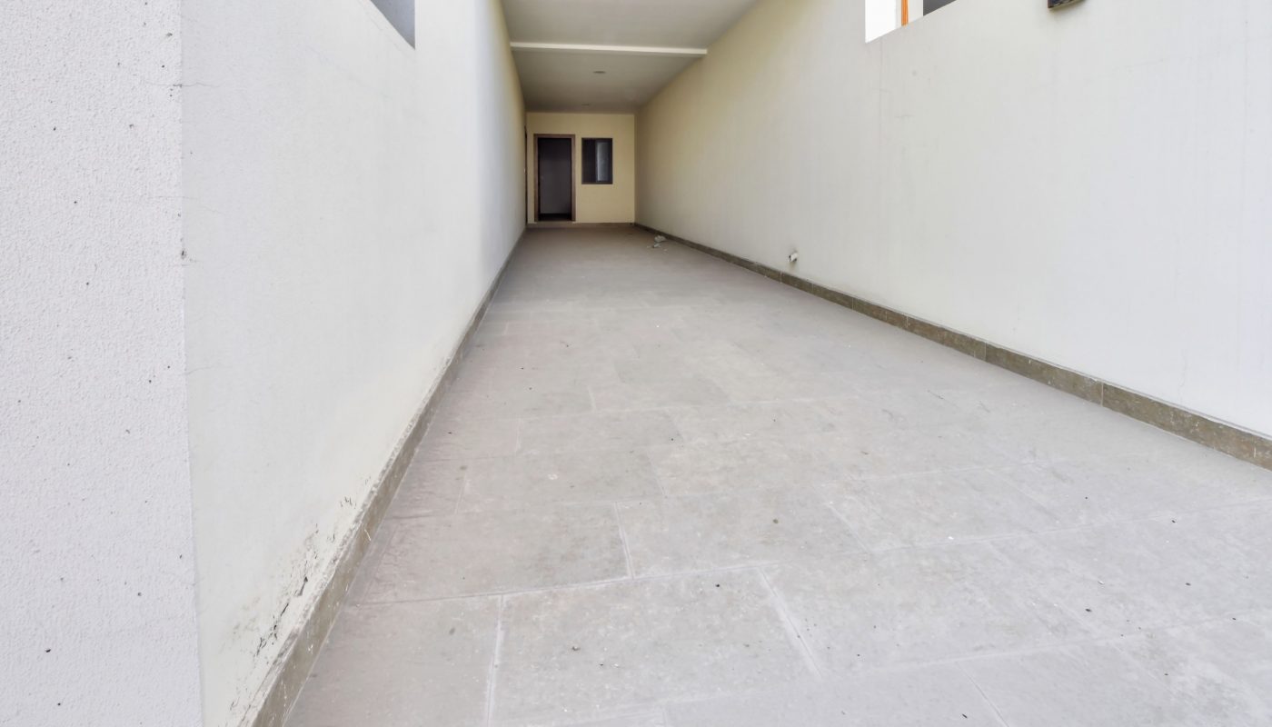 Long, narrow, covered walkway with white walls and a tiled floor leading to a dark doorway at the end.