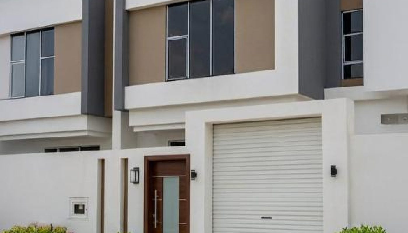 Modern two-story house with large windows, a wooden front door, and a white roll-up garage door. Minimalist landscaping with bushes and grass lines the entrance.