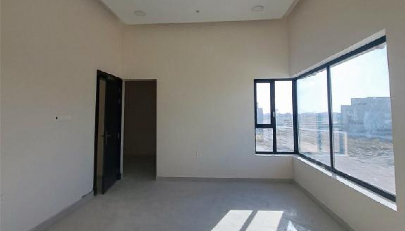An empty, unfurnished room with white walls, tiled floor, a black door, and large windows letting in sunlight.
