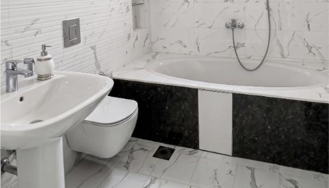 A modern bathroom with white marble tiles, a wall-mounted toilet, pedestal sink, soap dispenser, bathtub with showerhead, and a small window above the tub.