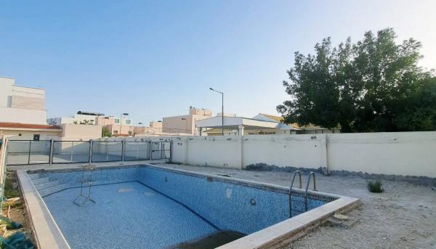 An empty rectangular swimming pool with ladders and safety railings, surrounded by gravel, in a fenced backyard with houses and a tree in the background under a clear sky.