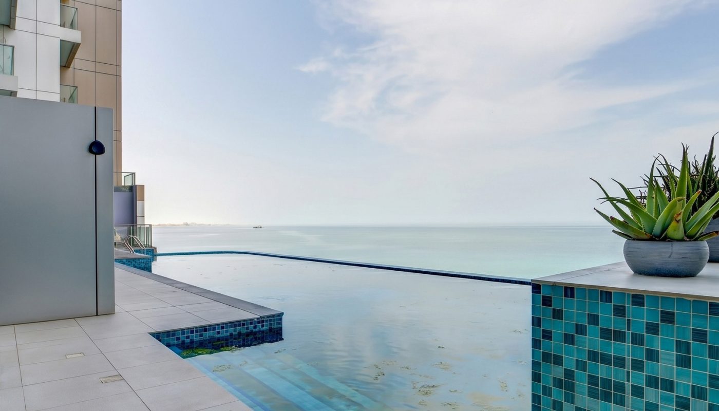 Modern rooftop infinity pool with blue tiles and a potted succulent overlooks a calm ocean under a partly cloudy sky.