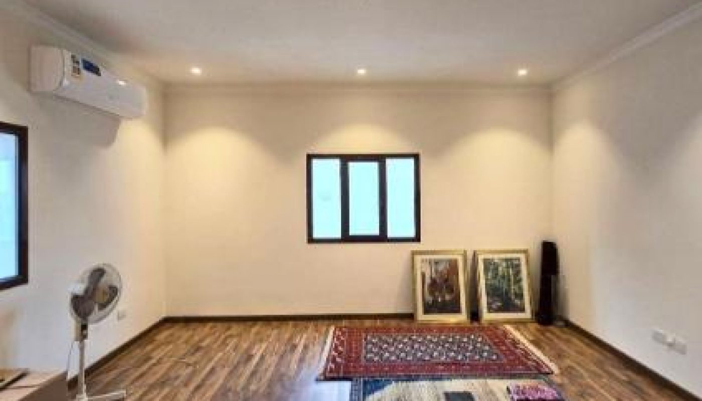 A nearly empty room with wooden floors, two rugs, three framed artworks on the floor, a fan, a box, and two windows. An air conditioner is mounted on the wall.