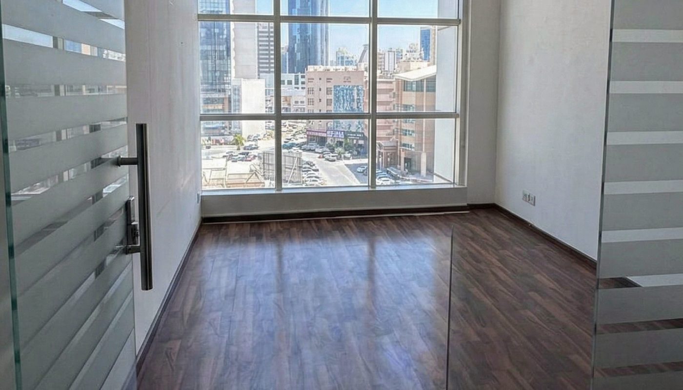 Empty office room with wooden flooring, glass door, and large windows showing city buildings outside in daylight.