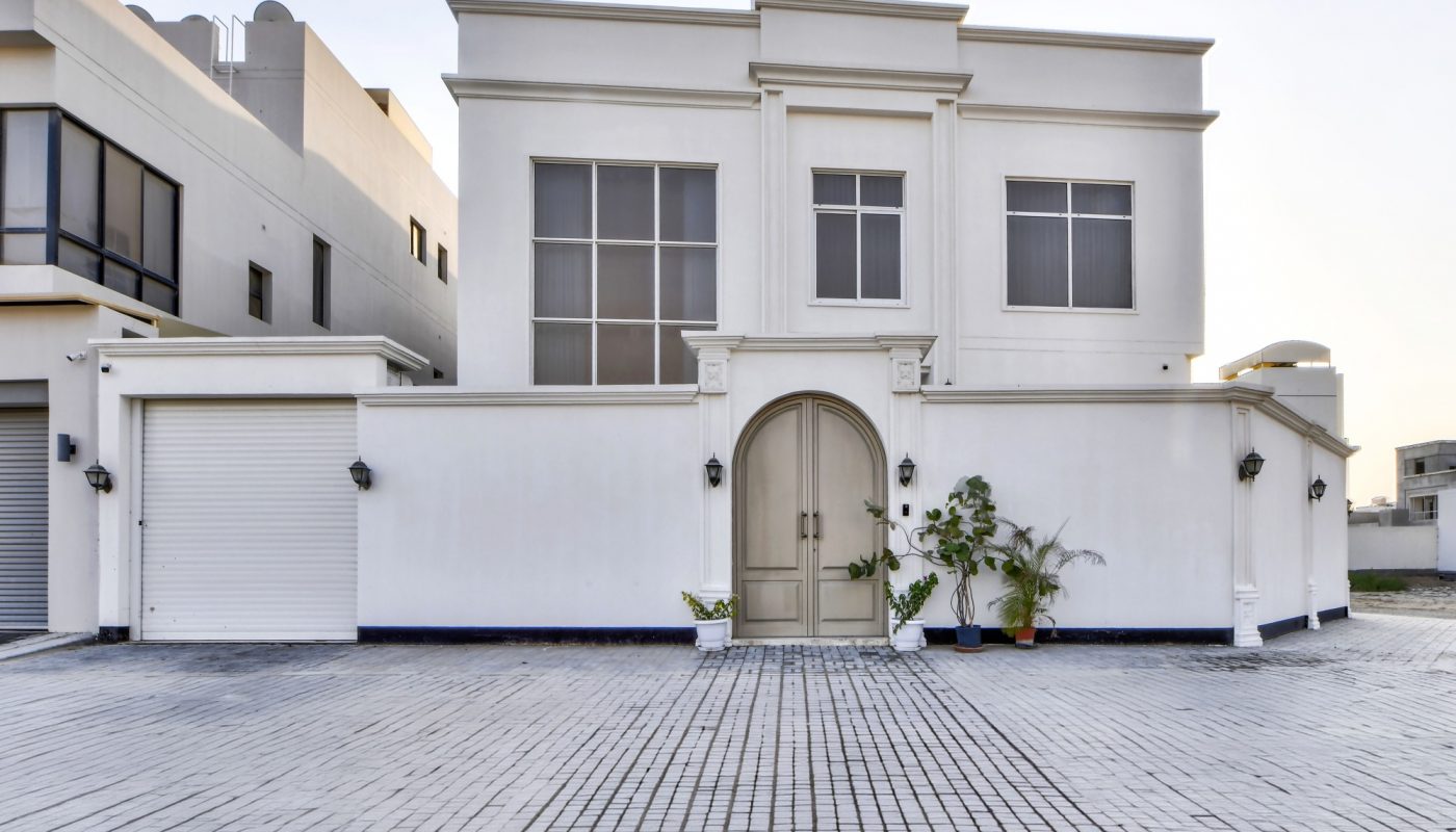Modern white house with arched double doors, large windows, garage door on the left, and potted plants by the entrance. Broad paved driveway in front.