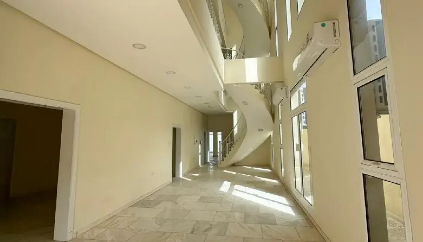 A modern, sunlit interior hallway with marble floors, high ceilings, tall windows, and a spiral staircase in the background.