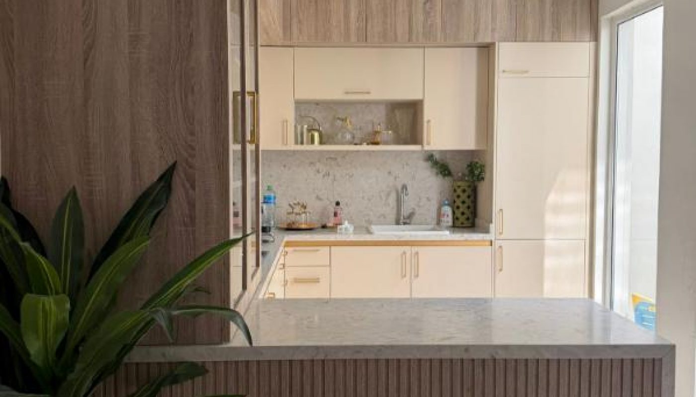 Modern kitchen with light wood cabinets, marble countertops, a potted plant, a stool with a gold frame, and sunlight coming through a glass door.