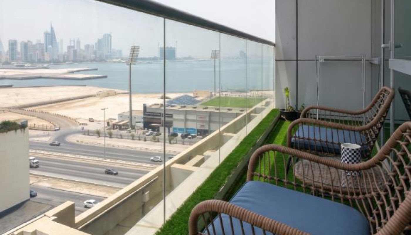 Balcony with two wicker chairs and green artificial grass, overlooking a city skyline, water, and a stadium through a glass railing.