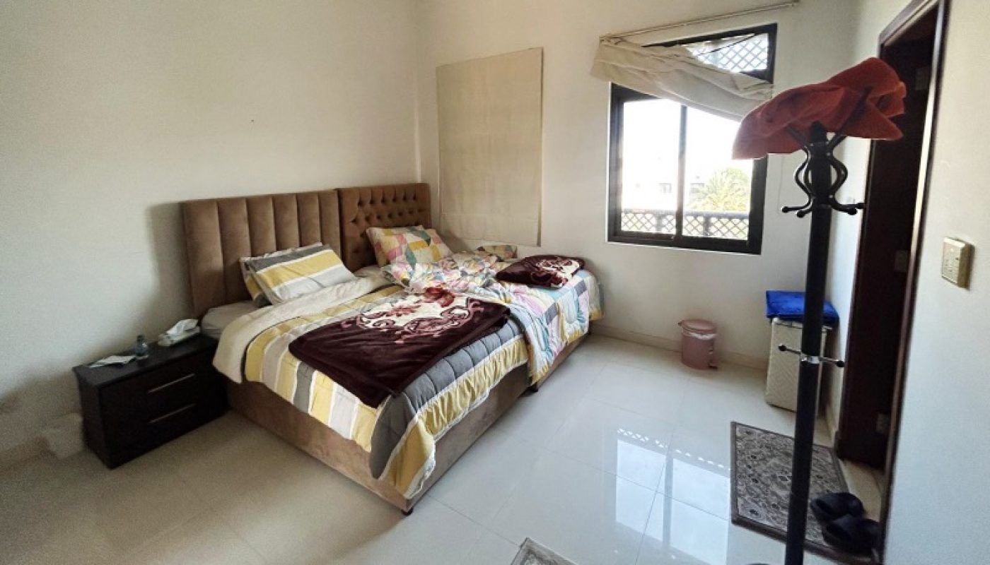 Bedroom with two single beds pushed together, a tufted headboard, nightstand, coat rack, window with open blinds, and light-colored tile floor with a rug.