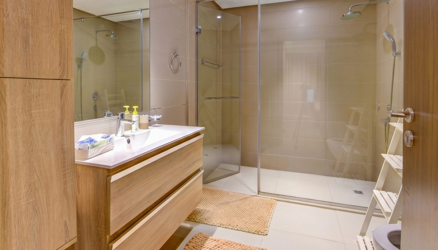 Modern bathroom with wood cabinets, double sink, two beige rugs, glass shower enclosure, and overhead lighting.