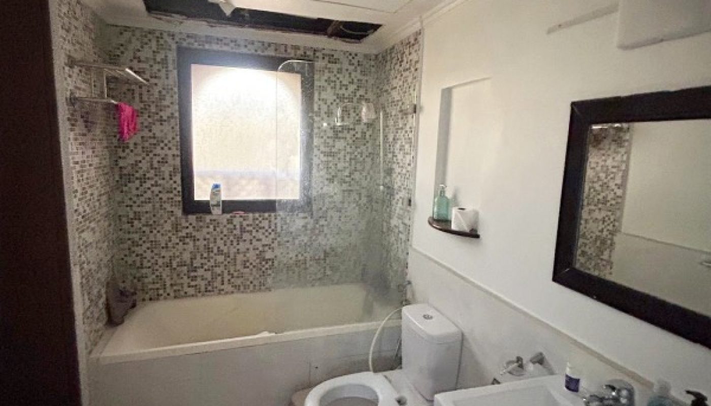 A small bathroom with a bathtub, toilet, sink, mirror, and toiletries, featuring white walls, mosaic tiles, and a damaged ceiling with exposed sections.