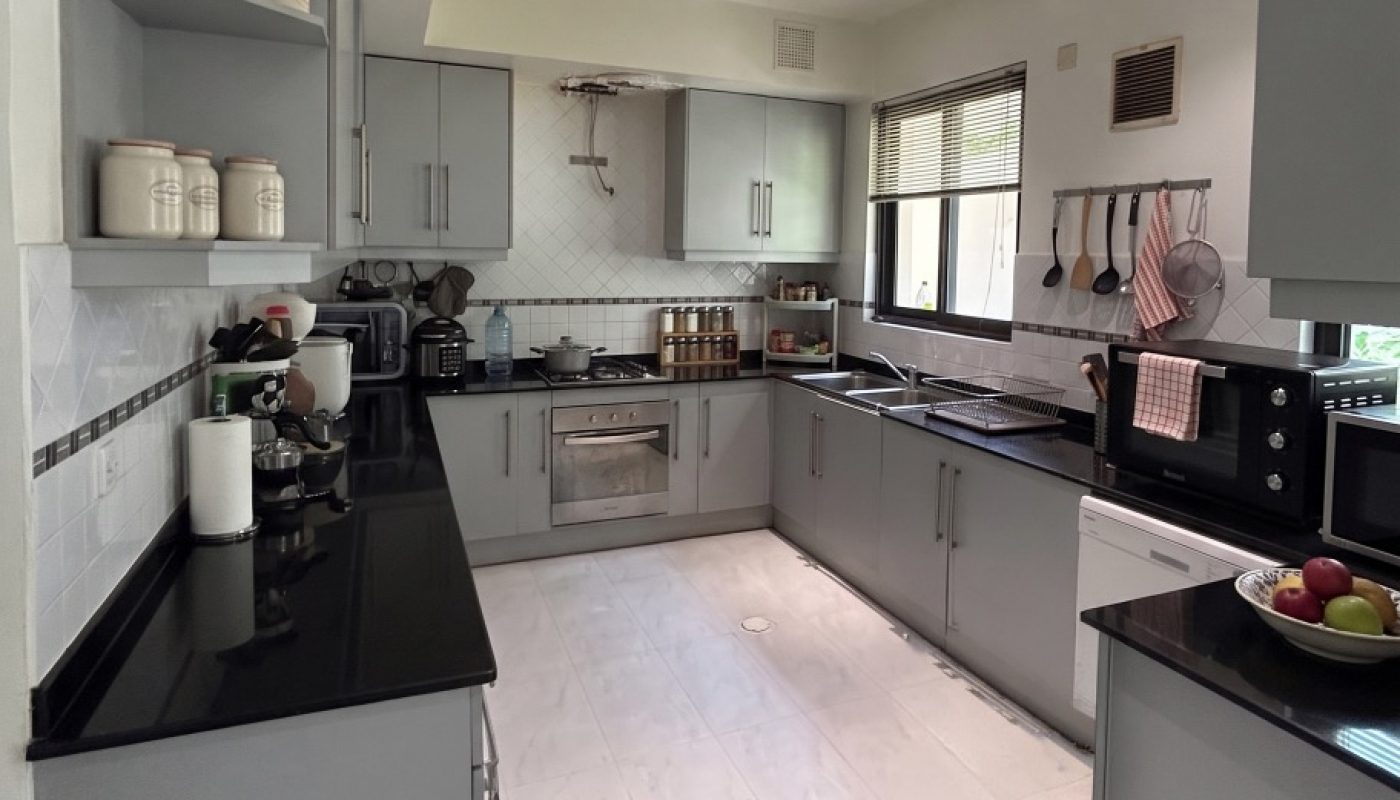 Modern kitchen with grey cabinets, black countertops, open shelves, white tile floor, and various appliances including a microwave, stove, kettle, and blender. Bowl of fruit on counter.