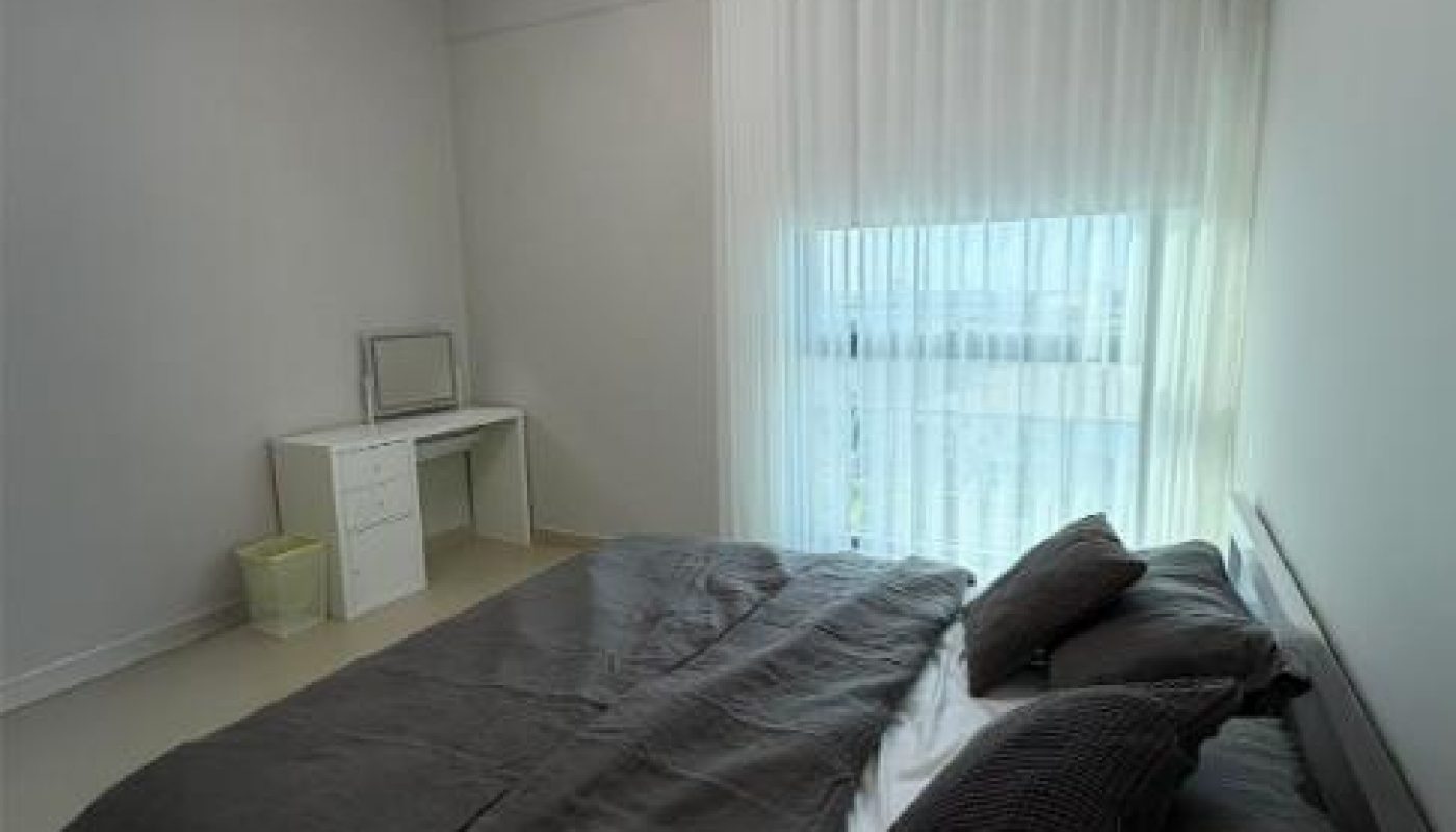 A minimal bedroom with a double bed covered in dark bedding, a small desk with a mirror, a wastebasket, sheer curtains, and a modern ceiling light.