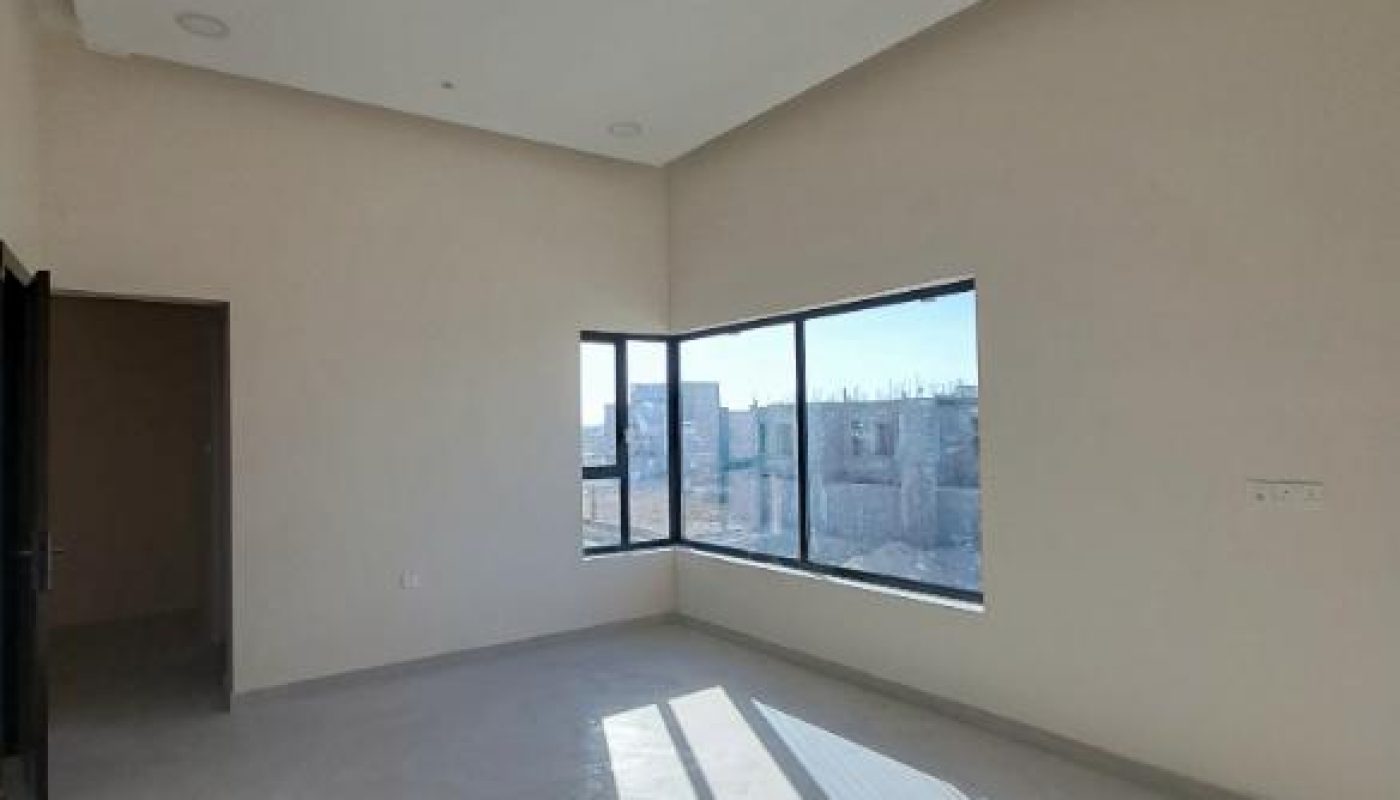 Empty, unfurnished room with light-colored walls, large windows letting in sunlight, and a door on the left side.