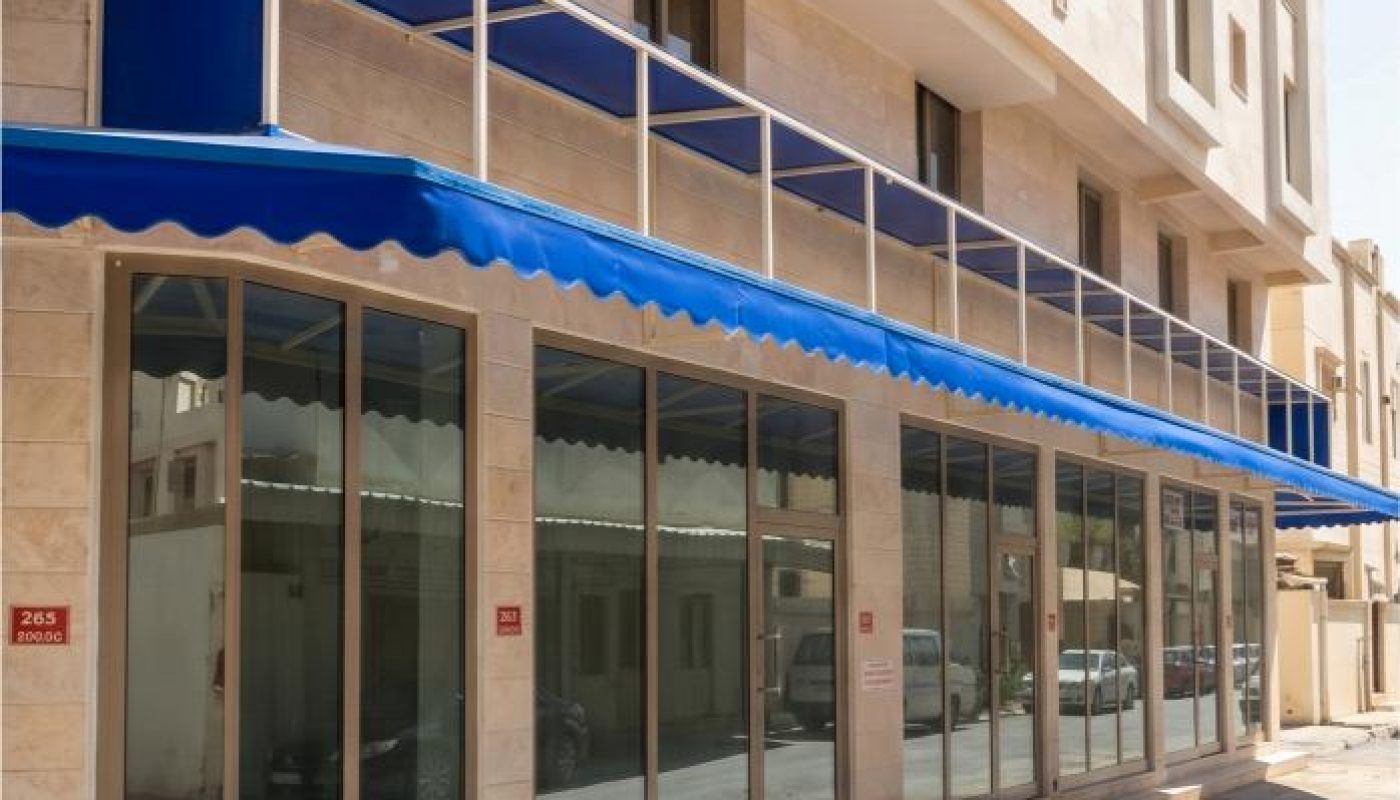 A modern building with large glass windows and a blue awning along the ground floor, located on a sunny, empty street.
