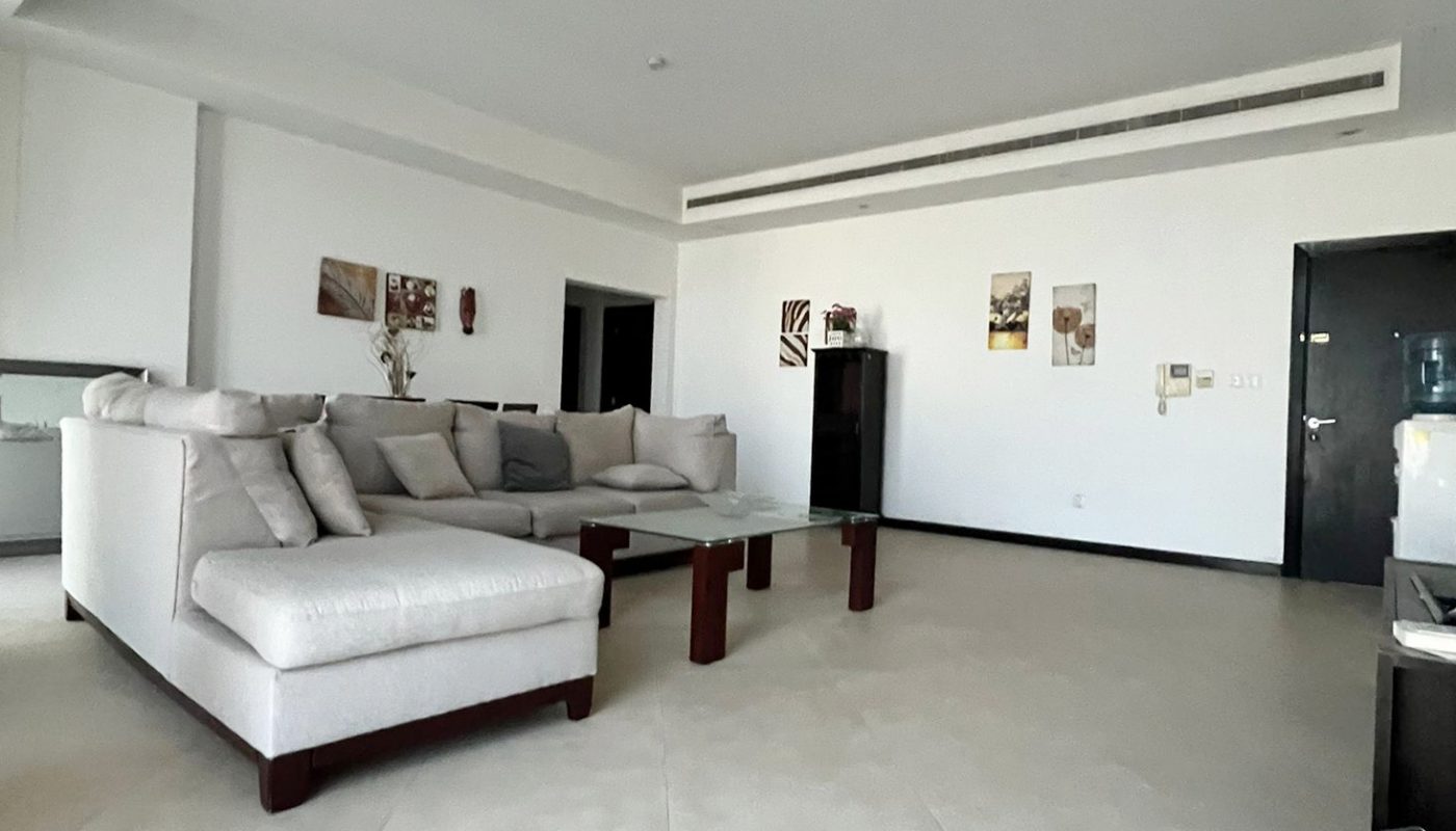 A spacious living room with a light sectional sofa, wooden coffee table, wall art, and light-colored tile flooring. The space is minimal and modern with white walls and a high ceiling.