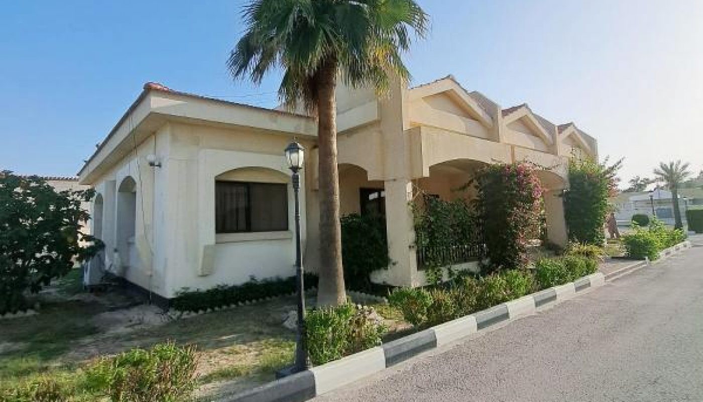 A single-story white house with a sloped roof, palm tree, and garden is situated next to a paved road under a clear blue sky.