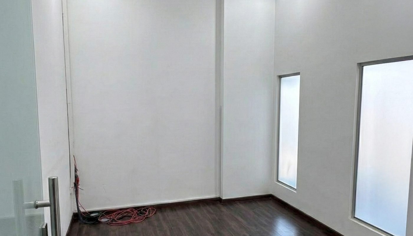 Empty room with dark wood flooring, white walls, high ceiling, two frosted windows, ceiling lights, and a coiled cable in the corner near the wall.