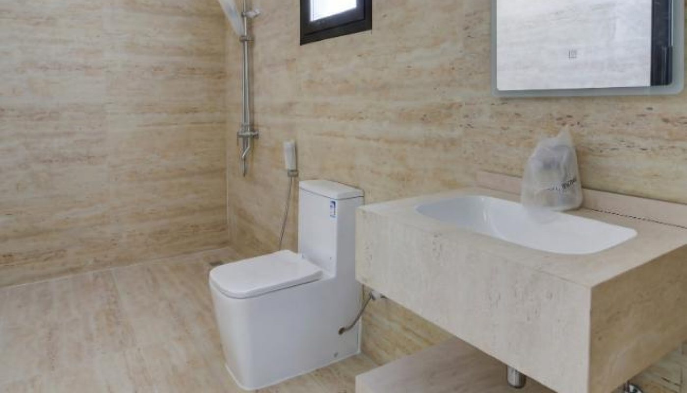 A modern bathroom with beige travertine tile walls and floors, featuring a white toilet, a wall-mounted sink with a stone vanity, and a walk-in shower.