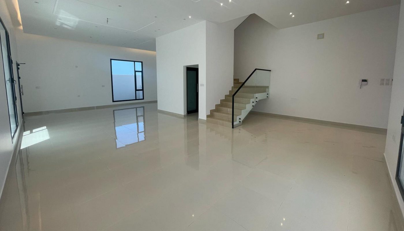 Spacious, empty modern room with glossy tiled floor, white walls, large windows, and a staircase with a glass railing leading to an upper level.